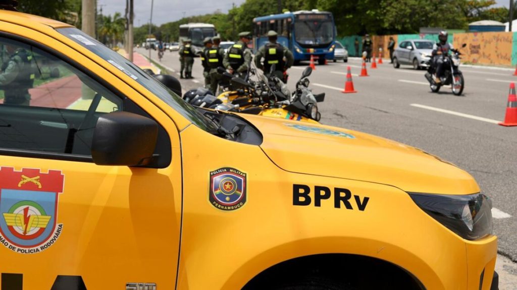 Foto mostra uma operação de trânsito realizada pelo Batalhão de Polícia Rodoviária (BPRv) de Pernambuco. Em primeiro plano, há uma viatura amarela da Polícia Militar com o brasão do BPRv e o logotipo da corporação. Atrás do veículo, aparecem policiais de uniforme verde com coletes reflexivos, além de motocicletas da polícia estacionadas. No fundo, há cones laranja delimitando a área da operação, um ônibus azul, alguns carros e motocicletas circulando na via. O céu está parcialmente nublado e há árvores e muros coloridos à beira da pista.
