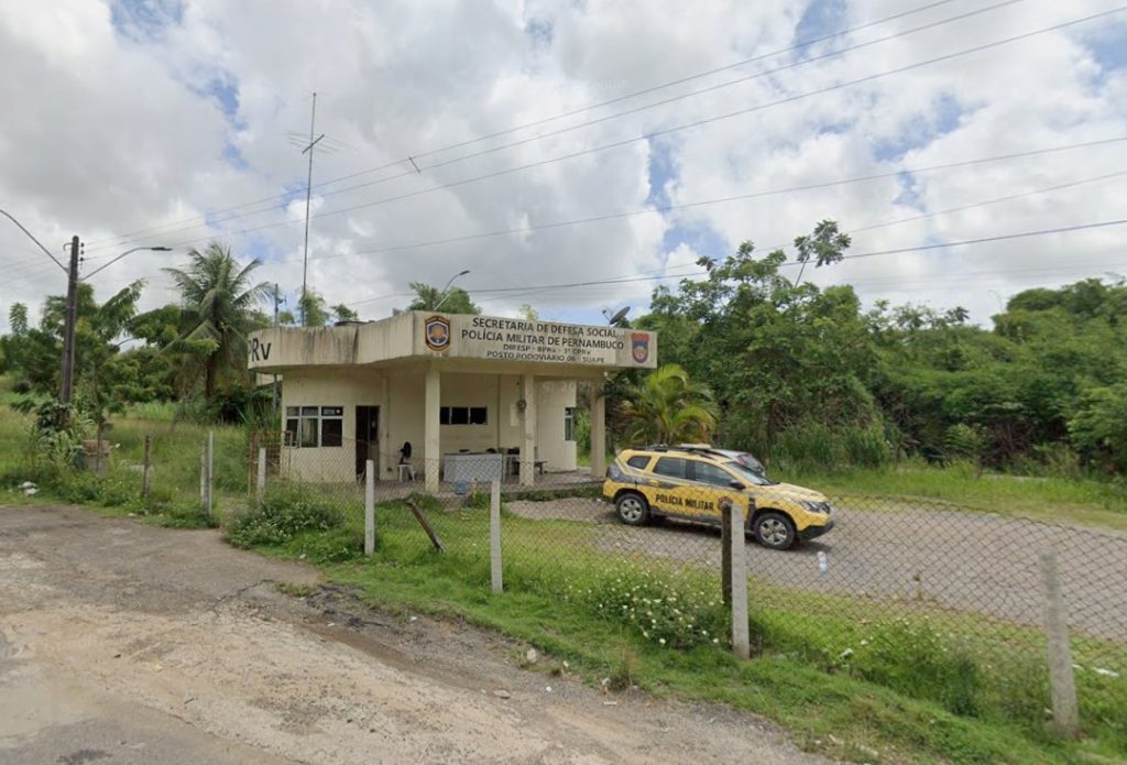 O ChatGPT disse: Foto mostra um pequeno posto da Polícia Militar de Pernambuco, localizado em área rural ou de beira de estrada, com vegetação densa e árvores ao fundo. O prédio é simples, de um pavimento, cor clara, com cobertura plana e um letreiro na fachada onde se lê “Secretaria de Defesa Social – Polícia Militar de Pernambuco – Posto Rodoviário 6º BPRv – Suape”. Em frente ao prédio há uma viatura amarela e azul da Polícia Militar estacionada. Um policial está sentado na varanda do posto. O local é cercado por uma cerca de arame e o céu está parcialmente nublado, com iluminação natural difusa.
