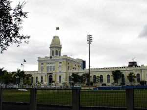 A foto mostra o quartel do comando da Polícia Militar de Pernambuco, um grande prédio histórico de cor amarela clara com detalhes brancos, em estilo neoclássico, localizado atrás de um campo gramado. A construção tem formato retangular, com uma torre central alta e arredondada no topo, onde tremula a bandeira do Brasil. Outras bandeiras estão hasteadas em mastros laterais. A fachada apresenta várias janelas altas em arco e ornamentos decorativos. Em frente ao edifício há árvores e postes de iluminação. O céu está nublado, criando uma luz difusa sobre a cena.
