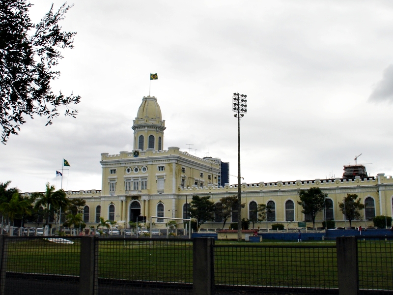 A foto mostra o quartel do comando da Polícia Militar de Pernambuco, um grande prédio histórico de cor amarela clara com detalhes brancos, em estilo neoclássico, localizado atrás de um campo gramado. A construção tem formato retangular, com uma torre central alta e arredondada no topo, onde tremula a bandeira do Brasil. Outras bandeiras estão hasteadas em mastros laterais. A fachada apresenta várias janelas altas em arco e ornamentos decorativos. Em frente ao edifício há árvores e postes de iluminação. O céu está nublado, criando uma luz difusa sobre a cena.