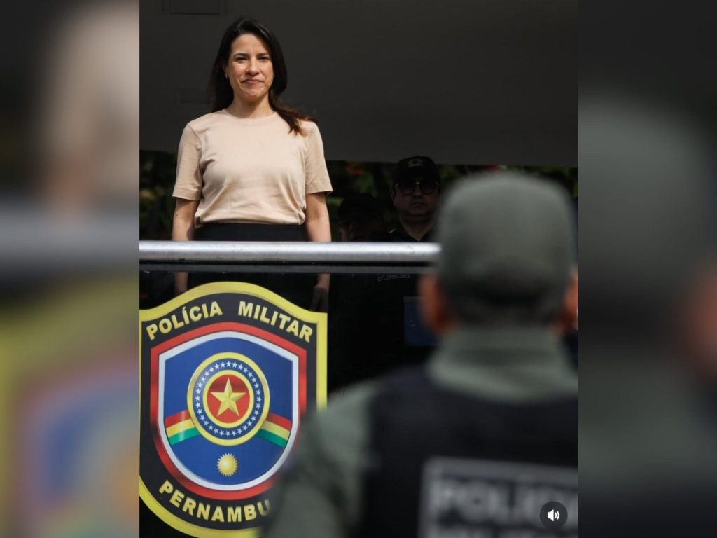 A foto mostra Raquel Lyra, mulher branca de cabelos escuros e lisos, de pé, em um palco ou palanque, com expressão serena e um leve sorriso. Ela veste uma camiseta bege clara e uma saia ou calça escura. À sua frente, há um policial de costas, usando uniforme verde e colete com a inscrição “Polícia Militar”. Na parte inferior da imagem, aparece o brasão da Polícia Militar de Pernambuco, com as cores azul, vermelha, amarela e verde. A cena ocorre em ambiente externo, com luz natural.