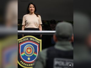 A foto mostra Raquel Lyra, mulher branca de cabelos escuros e lisos, de pé, em um palco ou palanque, com expressão serena e um leve sorriso. Ela veste uma camiseta bege clara e uma saia ou calça escura. À sua frente, há um policial de costas, usando uniforme verde e colete com a inscrição “Polícia Militar”. Na parte inferior da imagem, aparece o brasão da Polícia Militar de Pernambuco, com as cores azul, vermelha, amarela e verde. A cena ocorre em ambiente externo, com luz natural.