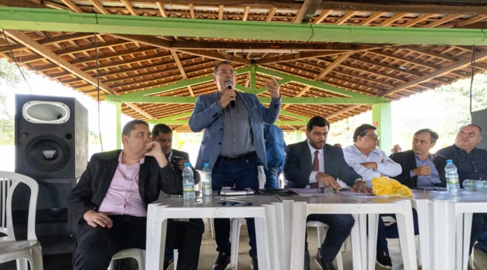 A foto mostra uma reunião em um espaço coberto com telhado de madeira e colunas verdes. Sete homens estão sentados atrás de duas mesas plásticas brancas, dispostas lado a lado. Um deles, de pé no centro, segura um microfone e fala ao público — ele veste calça jeans, camisa azul e paletó cinza-claro. Os demais o observam; alguns estão com expressão séria, outros atentos. Há garrafas de água, papéis e um microfone sobre as mesas. À esquerda, há uma grande caixa de som preta. O ambiente é simples e ventilado, com iluminação natural. A cena sugere uma reunião ou audiência pública.