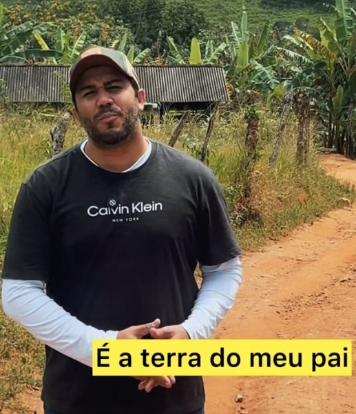 A foto mostra um homem em pé em uma estrada de terra cercada por vegetação. Ele tem barba curta, usa boné e veste uma camiseta preta da marca Calvin Klein sobre uma blusa de manga comprida cinza-clara. Atrás dele há uma cerca de arame e, mais ao fundo, uma casa simples com telhado de amianto, cercada por bananeiras e árvores. O cenário é rural, com morros e muito verde ao redor. Na parte inferior da imagem, aparece uma faixa amarela com o texto: “É a terra do meu pai”.
