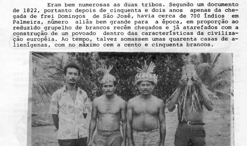A imagem em preto e branco mostra quatro homens em pé, lado a lado, posando para a foto em um ambiente externo com vegetação ao fundo. Três deles estão sem camisa e usam cocares ou adornos de palha na cabeça; o quarto homem, à esquerda, veste uma camisa de mangas curtas. Um dos homens cobre parcialmente o rosto com um adorno feito de folhas ou fibras. Acima da foto, há um trecho de texto impresso em português que menciona o número de indígenas e colonos brancos na região de Palmeira por volta de 1822, destacando a presença de cerca de 700 indígenas.