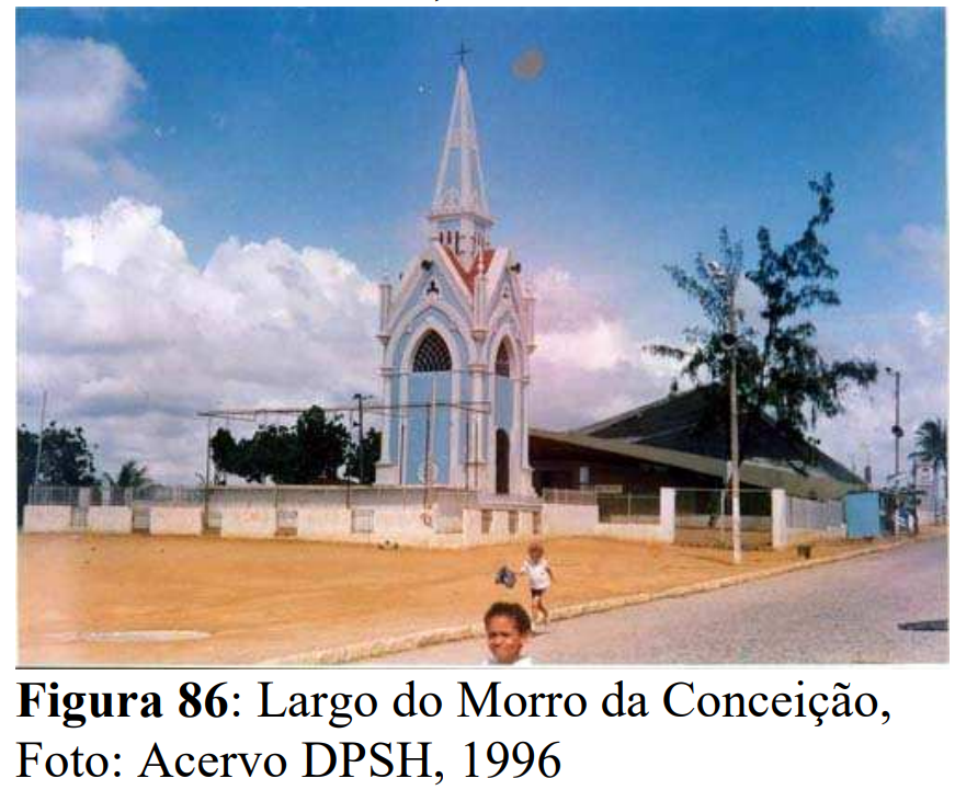 A foto mostra o Largo do Morro da Conceição, no Recife, em 1996. Em destaque, no centro da imagem, está o monumento da torre pontiaguda de cor bege, com detalhes brancos e azuis nas molduras e janelas em arco, típico do estilo neogótico. O monumento fica em uma área cercada por muro baixo e grade branca. À direita, há uma construção ampla com telhado escuro e muros claros. Na frente do largo, o chão é de terra batida e uma rua pavimentada passa ao lado. Duas crianças caminham próximas ao monumento, e o céu está azul com nuvens brancas espalhadas, indicando um dia ensolarado.