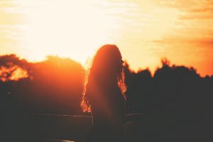 A imagem mostra a silhueta de uma pessoa de cabelos longos e soltos, vista de perfil, em pé ao ar livre durante o pôr do sol. O fundo é dominado por uma luz intensa e alaranjada do sol baixo no horizonte, que envolve toda a cena em tons quentes de dourado e laranja. A paisagem ao fundo tem árvores escuras, quase em sombra, e o céu exibe algumas nuvens dispersas iluminadas pela luz do entardecer.