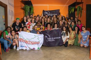 A imagem mostra um grupo diverso de pessoas reunidas em um espaço interno, posando para uma foto coletiva. Estão atrás de duas bandeiras: um da Rede de Proteção a Jornalistas e outro do Sindicato dos Jornalistas do Ceará (SINDJORCE). Algumas pessoas estão sentadas, outras em pé, todas sorrindo. O ambiente é acolhedor, com plantas penduradas e piso de azulejo. A cena transmite união e apoio à causa dos comunicadores.