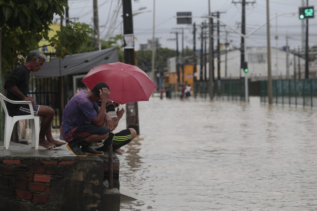 Na foto, uma rua está completamente alagada após uma forte chuva. Em primeiro plano, três homens observam a enchente. Um deles está sentado em uma cadeira plástica branca, enquanto os outros dois estão agachados na beira da calçada, um deles segurando um guarda-chuva vermelho. Todos parecem preocupados e observam a água barrenta que cobre a via. Ao fundo, vê-se a continuidade da rua inundada, postes de energia, cercas e algumas pessoas caminhando pela água. O céu está nublado, reforçando o cenário de tempo chuvoso