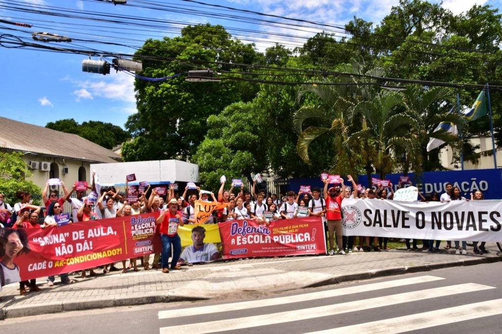 Um grupo grande de pessoas, formado por estudantes e educadores, participa de um protesto em frente a um prédio público identificado por uma placa azul com a palavra “Educação”. Os manifestantes ocupam a calçada e seguram faixas e cartazes coloridos. As faixas trazem mensagens como “Salve o Novaes”, “Defenda a escola pública” e “Sauna de aula, não dá! Cadê o ar-condicionado?”. Muitos participantes levantam os braços e demonstram entusiasmo. O céu está claro e há árvores verdes ao fundo, sugerindo um dia ensolarado.