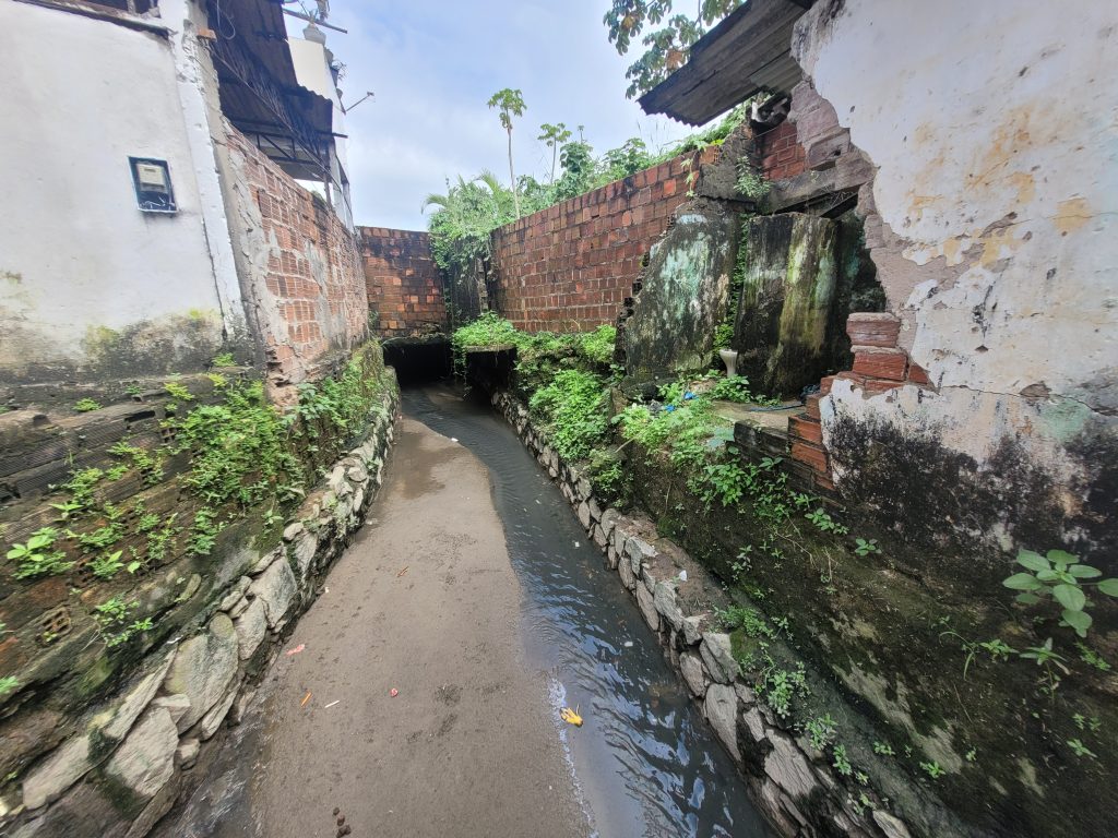 A foto mostra um canal estreito de esgoto a céu aberto, passando entre muros de casas simples construídas com tijolos aparentes e paredes desgastadas. As margens do canal são revestidas com pedras e há vegetação crescendo nos muros úmidos e nas bordas. À esquerda, uma parede branca apresenta manchas de mofo e rachaduras; à direita, uma parede está parcialmente destruída, revelando tijolos e infiltrações. Ao fundo, o canal segue em direção a um túnel escuro sob uma estrutura de alvenaria. O céu está nublado, e há algumas árvores e plantas visíveis acima dos muros.