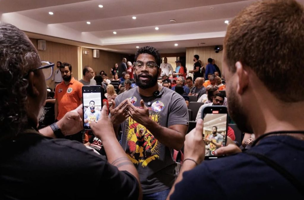 Na imagem, Jones Manoel é um homemnegro, de barna, que está em pé, falando para duas pessoas que o gravam com celulares em um auditório cheio. Ele usa óculos, camiseta cinza com estampa colorida e botons no peito. O ambiente é iluminado, com várias pessoas sentadas ao fundo, algumas usando roupas de cores fortes como laranja e azul. Jones aparece gesticulando enquanto fala, em um contexto que parece ser um encontro, reunião ou evento político.