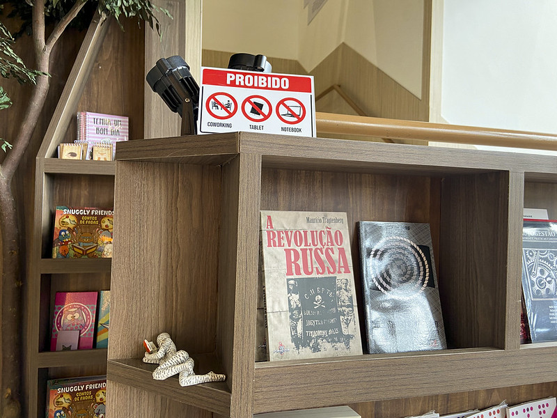 A foto mostra uma prateleira de madeira em uma livraria. No topo da estante há uma placa vermelha e branca com a palavra “PROIBIDO” em destaque, seguida de ícones que indicam ser proibido o uso de coworking, tablet e notebook. Ao lado da placa, há um par de binóculos. Na prateleira abaixo, estão expostos livros, entre eles “A Revolução Russa”, de Maurício Tragtenberg, e outros títulos de capa escura. À esquerda, parte de outra estante aparece com livros infantis coloridos.