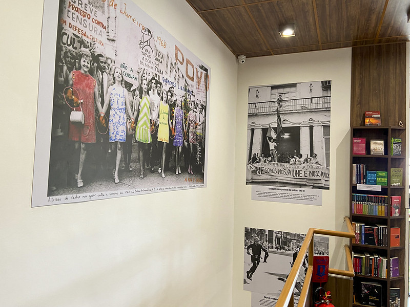 A foto mostra uma parede interna de uma livraria decorada com grandes painéis fotográficos em preto e branco que retratam momentos históricos de protestos no Brasil. Na parede à esquerda, há uma imagem de mulheres usando vestidos coloridos, marchando em um protesto no Rio de Janeiro em 1968 contra a censura. À direita, há duas fotos em preto e branco: a superior mostra pessoas em uma sacada com uma faixa escrita “A UNE SOMOS NÓS, A UNE É NOSSA VOZ”; a inferior exibe um manifestante correndo em meio a um protesto de rua. No canto direito da imagem, vê-se parte de uma estante com livros organizados por cor e uma escada de madeira que leva ao andar superior.