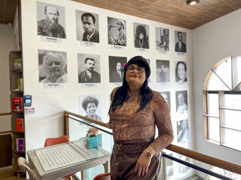 A foto mostra Rita de Cássia Valena, uma mulher em pé dentro de uma livraria ou café literário, posicionada no mezanino do espaço. Ela tem cabelos longos pretos e escuros, usa óculos, uma blusa rendada marrom e saia da mesma cor, com cinto xadrez. Usa relógio dourado e uma flor presa na boina preta sobre o cabelo. Atrás dela, uma parede exibe retratos em preto e branco de figuras conhecidas da literatura e do pensamento, organizados em duas fileiras, cada um com o nome impresso abaixo. À direita, uma grande janela em arco deixa entrar luz natural, iluminando o ambiente.