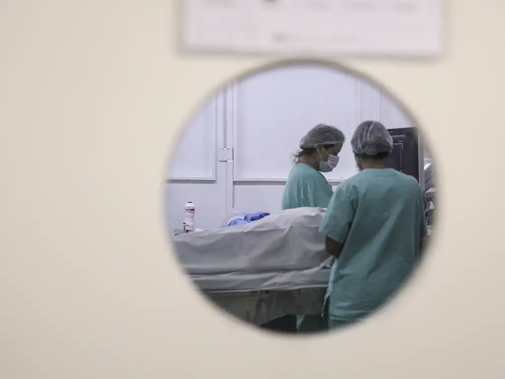 A foto mostra uma cena hospitalar vista através de uma pequena janela redonda em uma porta. No centro da imagem, aparecem duas pessoas com roupas cirúrgicas verdes, toucas descartáveis e máscaras de proteção. Elas estão ao lado de um leito hospitalar onde há um paciente coberto por um lençol branco. O ambiente tem paredes claras e equipamentos médicos ao fundo, transmitindo a ideia de uma sala de atendimento ou procedimento clínico.