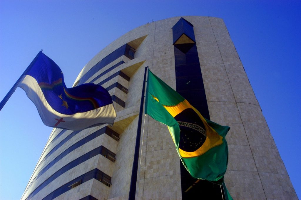 A foto mostra a fachada do prédio-sede do Tribunal Regional Federal da 5ª Região (TRF-5), em Recife. A imagem é feita de um ângulo baixo, destacando a arquitetura moderna do edifício, que tem formato curvo, revestimento em pedra clara e faixas horizontais de janelas escuras. À frente do prédio, duas bandeiras tremulam ao vento: à esquerda, a bandeira de Pernambuco, com fundo azul, arco-íris, sol, estrela e cruz vermelha; à direita, a bandeira do Brasil, verde com losango amarelo e círculo azul ao centro. O céu está limpo e azul intenso, compondo um contraste marcante com as cores das bandeiras e o tom claro da construção.