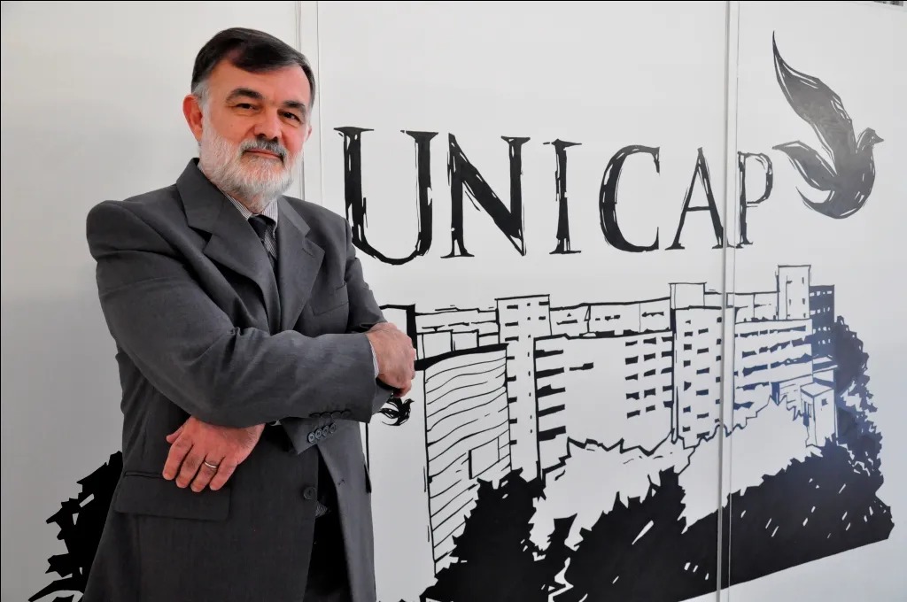 O ChatGPT disse: A foto mostra um homem de meia-idade em pé, posando diante de um painel ilustrado com o nome “UNICAP” e um desenho em preto e branco dos prédios da Universidade Católica de Pernambuco. Ele tem cabelo curto e barba grisalha, veste terno cinza escuro com gravata e camisa clara, e está com os braços cruzados, olhando para a câmera com expressão serena. À direita do nome “UNICAP”, há o desenho estilizado de uma pomba, símbolo frequentemente associado à instituição. O fundo branco e o contraste do desenho preto dão destaque à figura do homem em primeiro plano.