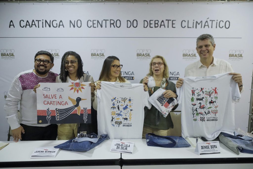 Cinco pessoas estão lado a lado atrás de uma mesa, posando para a foto em um evento. Elas seguram camisetas brancas estampadas com ilustrações coloridas e mensagens sobre preservação da Caatinga. A mesa à frente tem mais camisetas dobradas. Ao fundo, um painel branco traz a frase em destaque: “A Caatinga no centro do debate climático”. Todos sorriem e olham para a câmera, transmitindo um clima de celebração e engajamento ambiental.