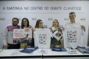 Cinco pessoas estão lado a lado atrás de uma mesa, posando para a foto em um evento. Elas seguram camisetas brancas estampadas com ilustrações coloridas e mensagens sobre preservação da Caatinga. A mesa à frente tem mais camisetas dobradas. Ao fundo, um painel branco traz a frase em destaque: “A Caatinga no centro do debate climático”. Todos sorriem e olham para a câmera, transmitindo um clima de celebração e engajamento ambiental.