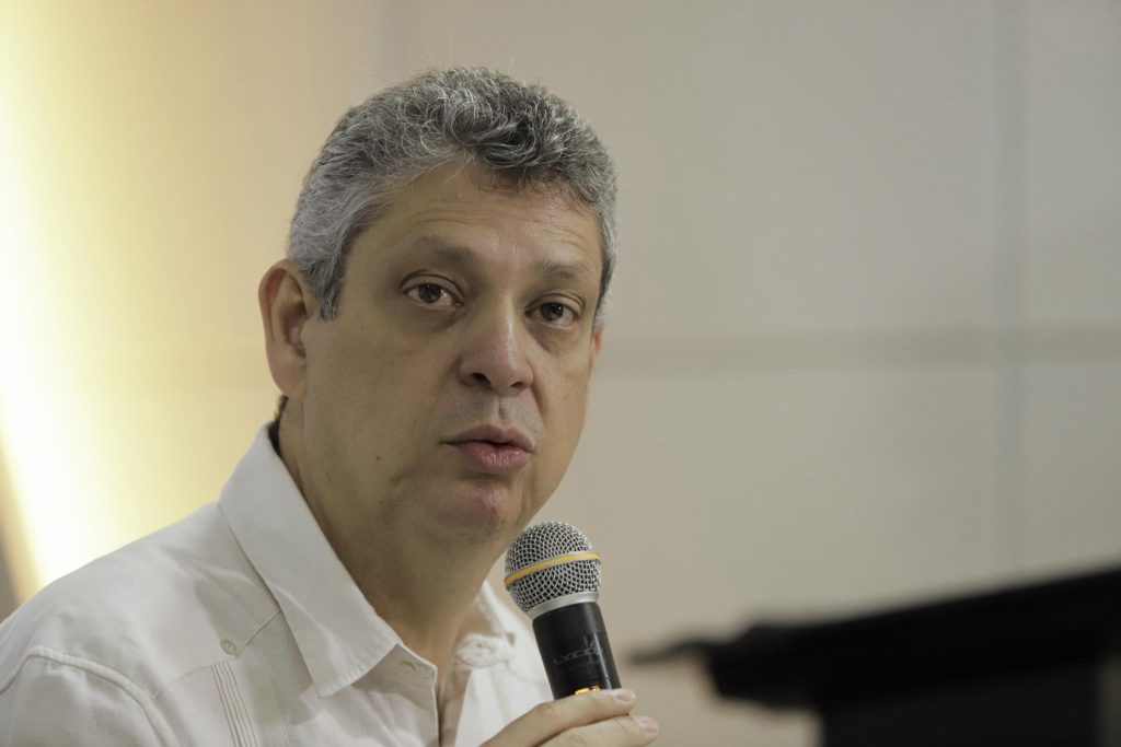 Foto do ministro Márcio Macêdo, um homem de pele clara e cabelos grisalhos, curtos e encaracolados, aparece em primeiro plano, sentado, segurando um microfone próximo à boca. Ele veste uma camisa social branca e olha em direção à câmera com expressão séria. O fundo é neutro, em tons claros, com parte de uma estrutura escura desfocada à direita da imagem.