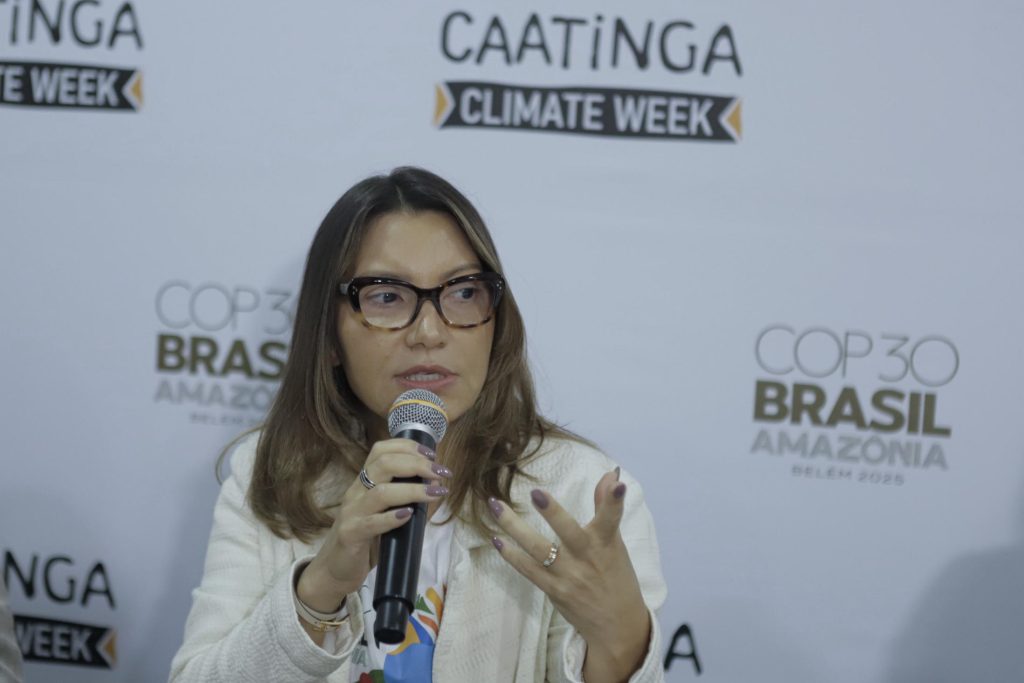 Foto da primeira-dama Janja, mulher de pele clara e cabelos castanhos lisos, na altura dos ombros, aparece em primeiro plano, sentada diante de um painel branco com os logos “Caatinga Climate Week” e “COP30 Brasil Amazônia”. Ela usa óculos de armação escura, casaco claro e segura um microfone na mão direita, enquanto gesticula com a esquerda. Olha para frente com expressão atenta, como se respondesse a uma pergunta ou fizesse uma intervenção no debate.