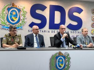 A foto mostra uma coletiva de imprensa na sede da Secretaria de Defesa Social de Pernambuco. Quatro homens estão sentados lado a lado atrás de uma mesa com microfones e gravadores. À esquerda, há um oficial da Polícia Militar com farda verde; os outros três usam terno e gravata. Um deles fala ao microfone, enquanto os demais escutam atentamente. Ao fundo, aparece uma parede com o grande logotipo azul “SDS” e o brasão do Governo de Pernambuco. O ambiente é formal e institucional.