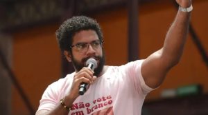 Foto de Jones Manoel, um homem de barba e cabelos crespos curtos fala ao microfone durante um evento ao ar livre. Ele usa óculos e uma camiseta clara com uma frase escrita em vermelho. O homem levanta um dos braços, como se enfatizasse o que está dizendo. Ao fundo, há uma estrutura de metal e uma parede de cor alaranjada desfocada.
