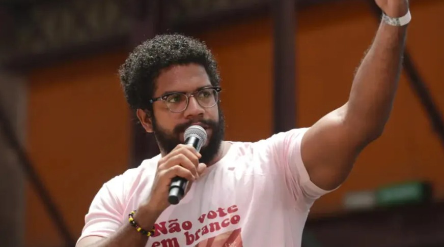 Foto de Jones Manoel, um homem de barba e cabelos crespos curtos fala ao microfone durante um evento ao ar livre. Ele usa óculos e uma camiseta clara com uma frase escrita em vermelho. O homem levanta um dos braços, como se enfatizasse o que está dizendo. Ao fundo, há uma estrutura de metal e uma parede de cor alaranjada desfocada.
