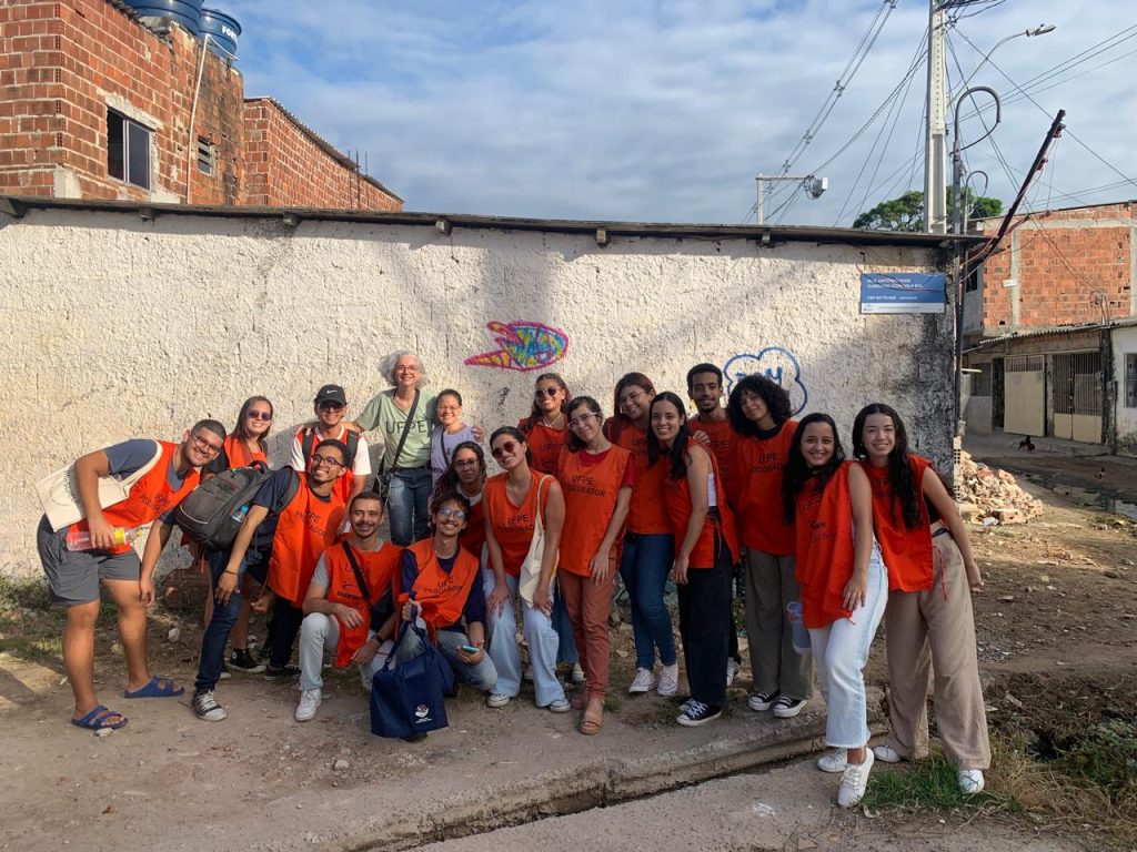 Um grupo de cerca de vinte pessoas posa sorridente em frente a um muro branco com pequenos grafites coloridos. A maioria veste coletes ou camisetas laranja com a inscrição “UFPE Extensionar”. Há pessoas de diferentes tons de pele e estilos, com expressões alegres e descontraídas. Algumas usam óculos de sol, outras carregam mochilas ou bolsas. O grupo está reunido em uma rua de bairro popular, com casas de tijolo aparente e muros simples ao redor. O céu está parcialmente nublado, e a luz do sol ilumina o grupo de forma suave, sugerindo fim de tarde.