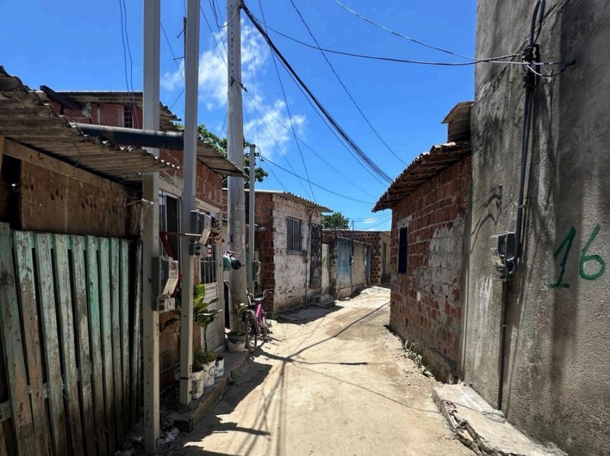 O ChatGPT disse: A foto mostra uma viela estreita em uma comunidade de casas simples, construídas com tijolos aparentes e coberturas de telha. O chão é de terra batida e há postes de energia elétrica com fios cruzando o céu. À esquerda, vê-se uma cerca de madeira verde-clara desgastada e, ao lado, uma pequena área com vasos de plantas e uma bicicleta encostada na parede. À direita, uma parede de concreto cinza tem o número “16” pintado em verde. O céu está limpo e azul, com algumas nuvens brancas, indicando um dia ensolarado.