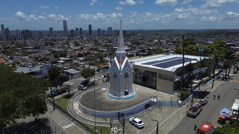 A imagem mostra uma vista aérea de um bairro urbano com muitas casas e alguns prédios altos ao fundo. Em destaque, no centro da foto, está um monumento bege com detalhes brancos e batentes azuis, em formato de torre pontiaguda que lembra uma pequena igreja ou capela gótica. O monumento fica em uma área circular cercada por um muro baixo e grades metálicas. Ao lado direito, há um prédio moderno de dois andares, com painéis solares no telhado e cercado por palmeiras. Em volta, é possível ver ruas com carros estacionados, algumas pessoas caminhando e barracas pequenas na calçada. O céu está azul com poucas nuvens e a iluminação indica que a foto foi tirada em um dia ensolarado.