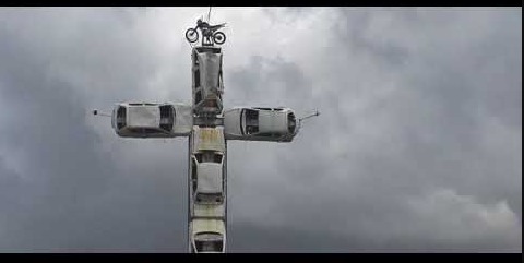 A imagem mostra uma escultura em forma de cruz feita com carros empilhados. São automóveis brancos e prateados colocados uns sobre os outros, formando o corpo e os braços da cruz. No topo da estrutura, há uma motocicleta posicionada de forma destacada. O céu ao fundo está nublado e cinzento, criando um clima dramático e contrastando com as cores claras dos veículos.