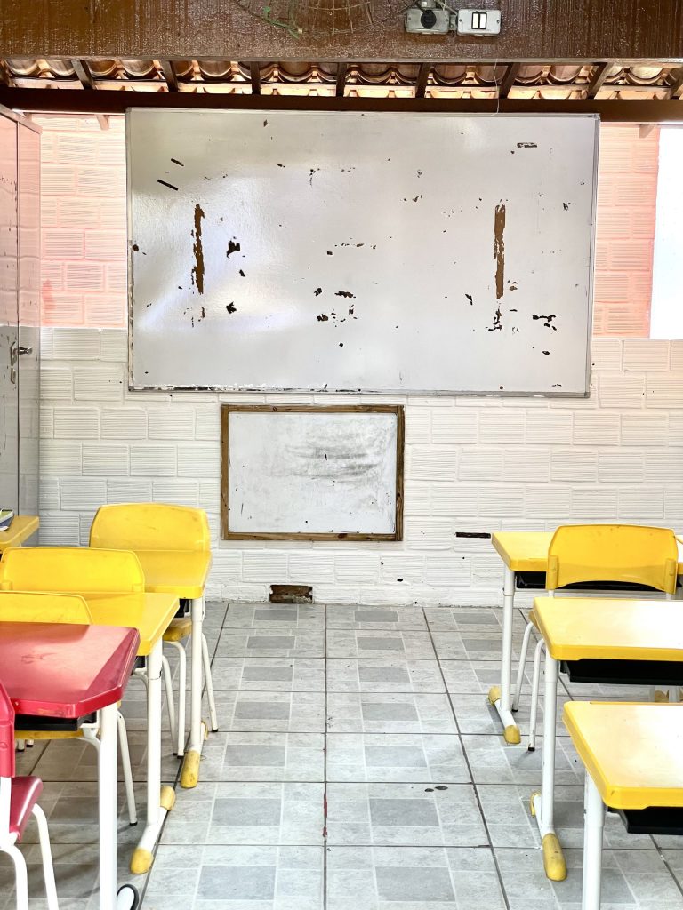 A foto mostra uma sala de aula simples de uma escola pública do Recife. No centro da imagem há um quadro branco grande, bastante desgastado, com partes descascadas que revelam o fundo marrom da parede. Abaixo dele, há um quadro menor, também gasto e emoldurado por madeira. O piso é de cerâmica clara com rejuntes escuros, e as paredes são revestidas com azulejos brancos em relevo. Na frente da sala, há carteiras individuais amarelas e uma vermelha, com cadeiras de plástico e pés de metal branco. A iluminação natural entra pelas laterais superiores, e o teto de telha cerâmica aparente é sustentado por vigas de madeira. A cena transmite a aparência de uma escola com poucos recursos e móveis bem usados.