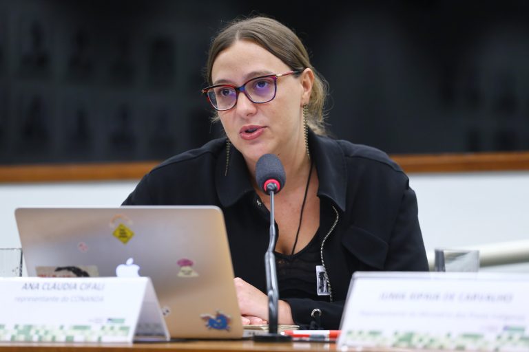 A foto mostra uma mulher sentada à mesa, falando ao microfone durante uma reunião ou audiência pública. Ela tem pele clara, cabelo castanho preso e usa óculos de armação roxa. Está vestida com uma blusa preta e usa brincos compridos. À sua frente há um notebook prateado com adesivos coloridos e uma placa de identificação com o nome “Ana Cláudia Cifali” e a informação de que é representante do Conanda. A mesa tem copos de água e papéis, e ao fundo há uma parede escura com quadros emoldurados desfocados. A imagem foi feita em um ambiente formal, possivelmente uma sala de comissões da Câmara dos Deputados