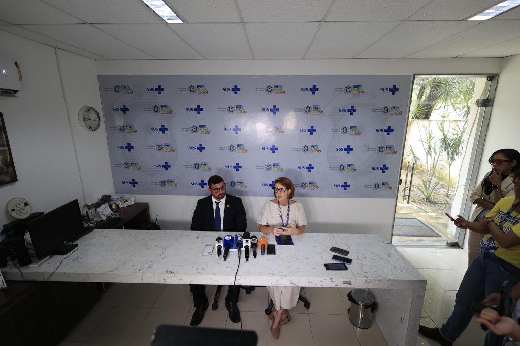 A foto mostra uma coletiva de imprensa em uma sala clara e simples. No centro, atrás de uma mesa de mármore, estão duas pessoas sentadas. À esquerda, um homem de barba, cabelo curto e óculos de grau. Ele veste terno preto, camisa branca e gravata escura. Ao lado dele, à direita, está uma mulher de cabelos curtos, castanhos claros. Ela usa óculos, veste roupa clara (blusa e calça brancas) e sandálias. Um crachá azul está pendurado em seu pescoço. Sobre a mesa, há microfones de imprensa, celulares e um caderno. Atrás deles, a parede tem logotipos do SUS e da Secretaria de Saúde de Pernambuco. À direita, três pessoas em pé observam e registram a coletiva com celulares.