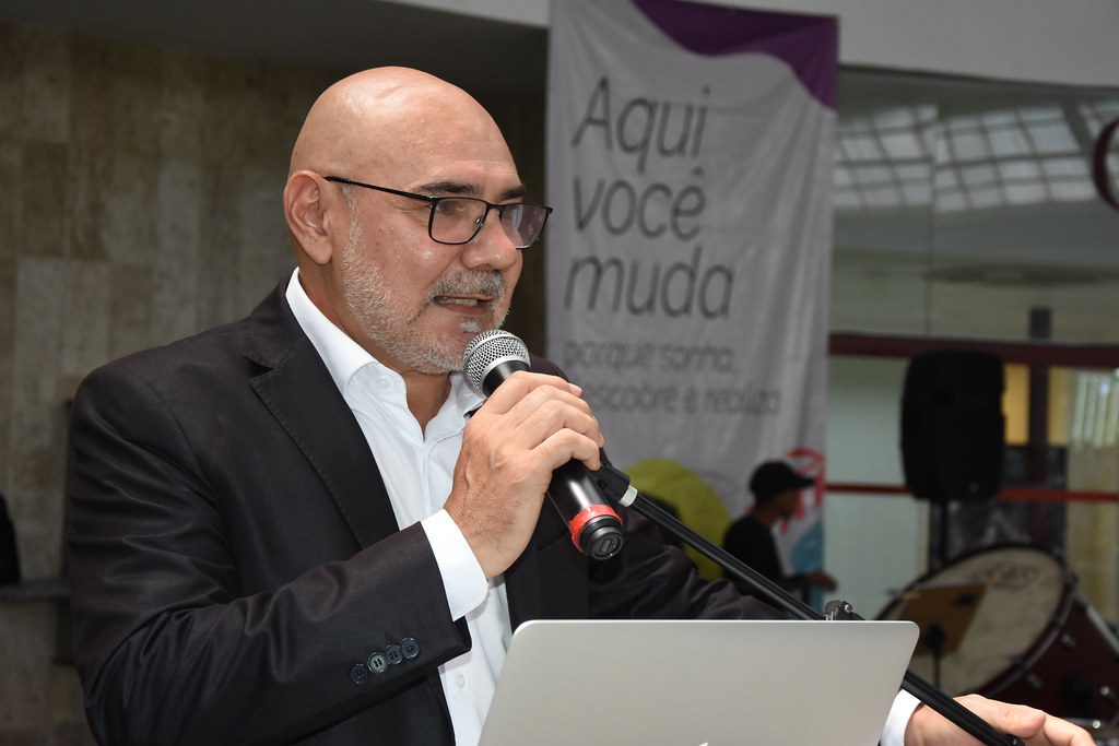 A foto mostra o reitor Alfredo Gomes, um homem calvo, de barba grisalha curta e óculos, falando ao microfone enquanto segura um notebook aberto. Ele veste terno escuro e camisa branca, sem gravata. Está em um ambiente interno, com parede de mármore claro ao fundo e um banner parcialmente visível com o texto “Aqui você muda porque sonha, se esforça e realiza”. Atrás dele, há uma pessoa desfocada e um instrumento musical grande, possivelmente um bumbo. A iluminação é clara, e o clima parece ser o de um evento formal ou institucional.