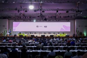 A imagem mostra um auditório amplo e bem iluminado, onde ocorre uma conferência internacional de alto nível. No palco, uma longa mesa acomoda diversos palestrantes sentados, com microfones e nomeações à frente, sugerindo a presença de delegações oficiais. Acima deles, uma tela digital exibe o título “UN Climate Change Conference – COP30 BRASIL – Belém - PA - 2025”, indicando que se trata da 30ª Conferência das Nações Unidas sobre Mudanças Climáticas. A plateia está organizada em fileiras, com participantes atentos e equipamentos de tradução simultânea, reforçando o caráter diplomático do evento