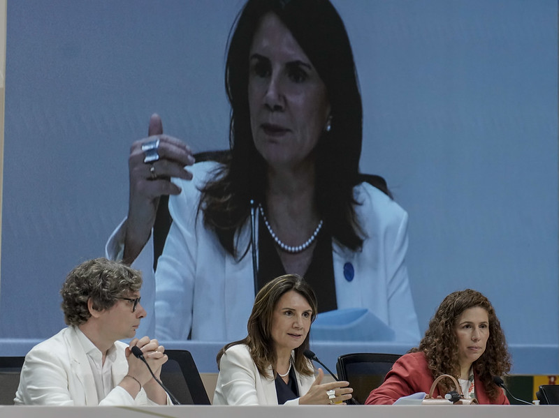 A imagem mostra três pessoas sentadas lado a lado em uma mesa de conferência, participando de um painel formal. À esquerda, um homem de óculos e cabelos grisalhos veste um blazer branco; ao centro, Ana Toni, uma mulher de cabelos castanhos escuros usa blazer branco e colar de pérolas, falando com gestos expressivos; à direita, outra mulher de cabelos cacheados veste blazer vermelho e observa atentamente. Ao fundo, uma grande tela exibe em destaque o rosto de Ana Toni, ampliando sua presença e reforçando seu papel de liderança na discussão. O ambiente é sério e profissional, com microfones e papéis sobre a mesa. A composição sugere um evento internacional, possivelmente ligado a temas políticos ou ambientais. A iluminação é clara e uniforme, favorecendo a visibilidade.