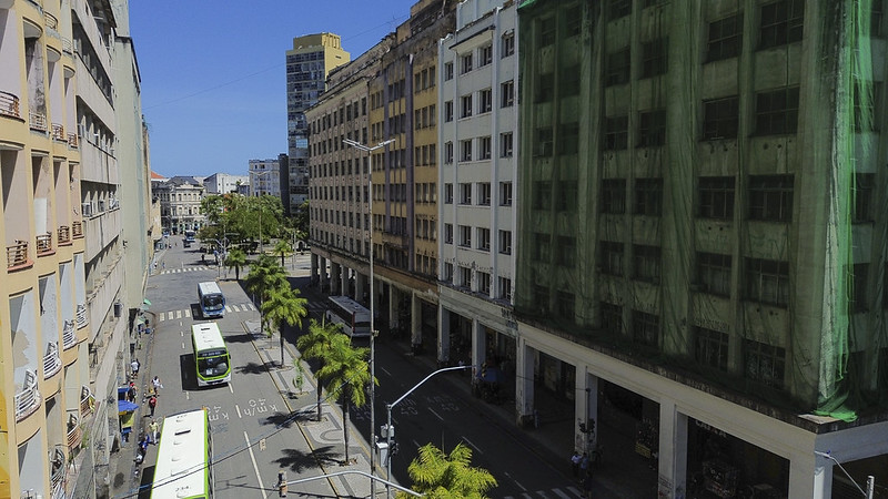 A imagem mostra uma vista aérea parcial da Avenida Guararapes em um dia ensolarado, sob céu azul. A avenida é larga e ladeada por prédios altos e antigos. À direita, um edifício grande está coberto por uma tela verde de proteção, indicando obra ou restauro. À esquerda, vê-se uma sequência de prédios com fachadas claras e varandas pequenas. A via conta com faixas exclusivas para ônibus, e alguns coletivos verdes circulam pela avenida. Há palmeiras altas plantadas no canteiro central, trazendo um toque de verde ao cenário urbano. Mais ao fundo, nota-se a continuação da avenida e outros prédios do centro do Recife, além de trechos de céu e algumas árvores compondo o horizonte.