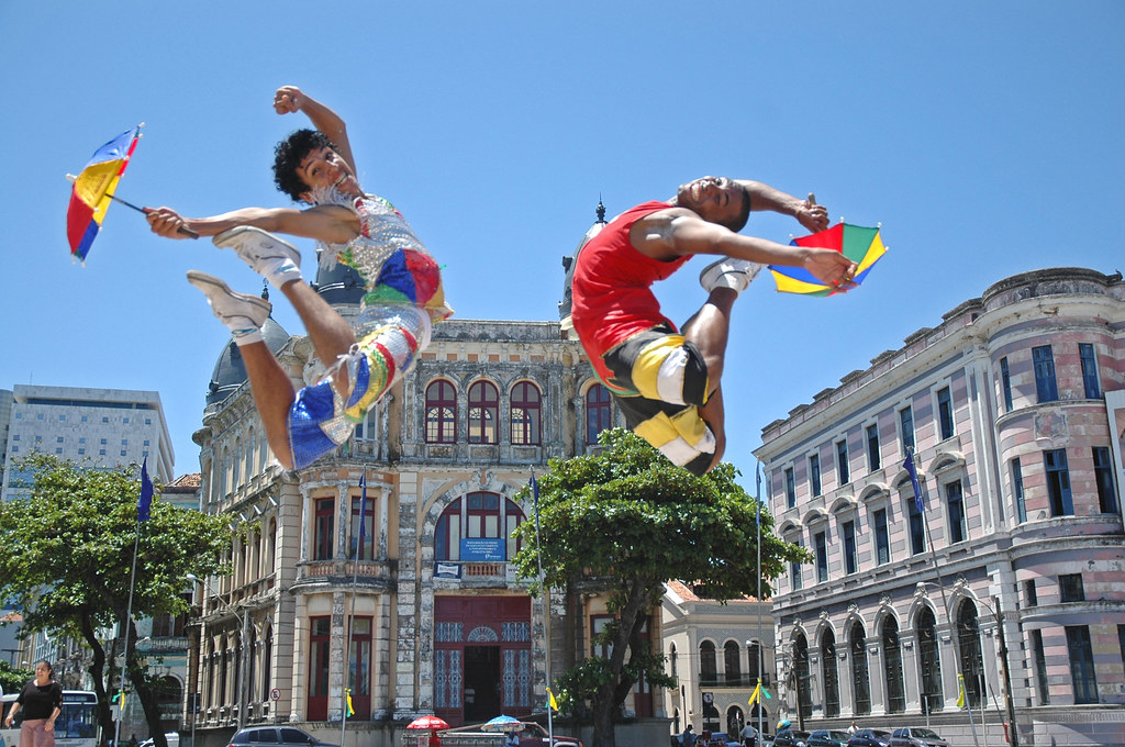 Dois passistas, caracterizados com roupas e sombrinha de frevo, saltando. Ao fundo, imagem de prédios do entorno da praça do Marco Zero, No Recife