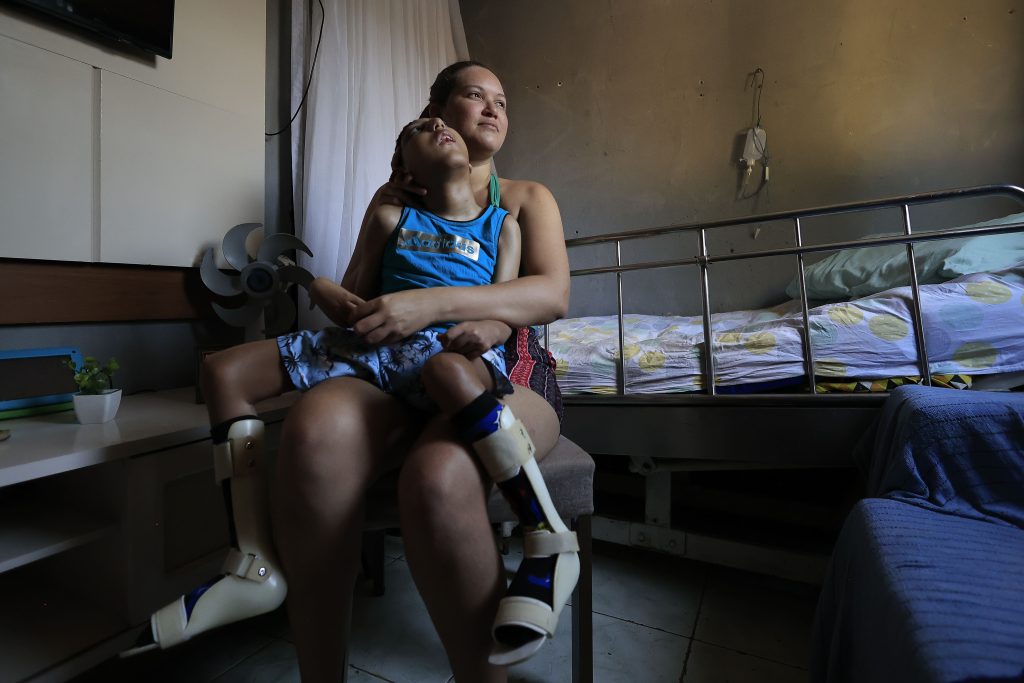 A imagem mostra uma mulher sentada na sala de uma casa simples, segurando no colo uma criança com microcefalia que usa órteses nas pernas. A mulher olha para frente com uma expressão protetora, enquanto a criança está apoiada em seu peito, olhando para cima. Ao lado, há uma cama hospitalar com lençóis coloridos e uma parede cinza ao fundo. O ambiente é modesto, mas transmite um sentimento de carinho, cuidado e vínculo entre mãe e filho.