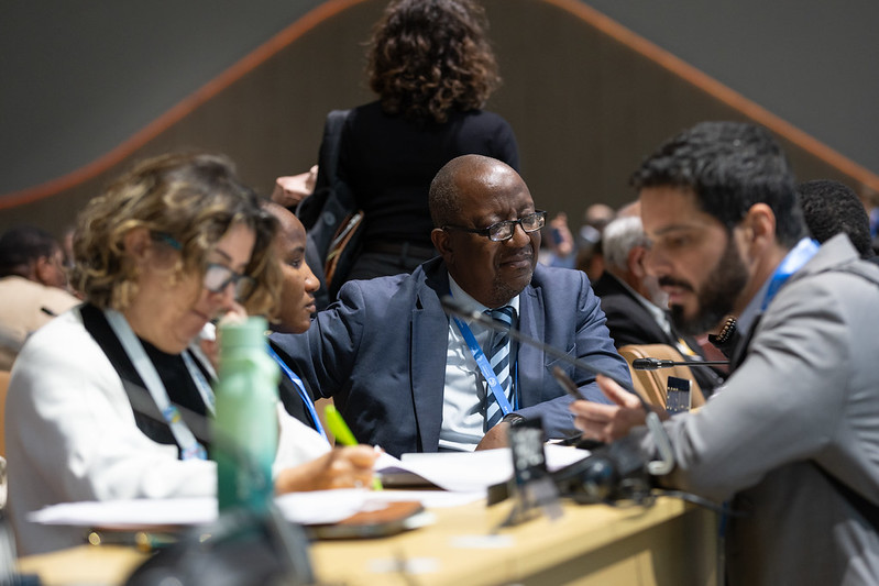 A foto mostra um grupo de pessoas reunidas em uma mesa durante uma conferência. No centro, um homem de terno azul e óculos conversa com outros participantes, enquanto segura alguns papéis. À sua esquerda, uma mulher escreve em um caderno, e à direita, um homem de barba lê um documento. Todos parecem concentrados na discussão. Ao fundo, há outras pessoas sentadas, também envolvidas em conversas. O ambiente é iluminado e tem aparência formal, típico de um encontro internacional ou evento diplomático.