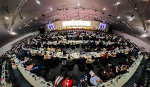 A foto mostra a abertura da COP30, realizada em um amplo auditório moderno e bem iluminado. O espaço está repleto de mesas e cadeiras organizadas em fileiras, todas ocupadas por participantes atentos. No fundo da sala, há um palco extenso onde vários palestrantes estão sentados atrás de uma longa mesa, participando de um painel de discussão. Telões grandes posicionados nas laterais do palco exibem os rostos dos palestrantes, facilitando a visualização para todos os presentes. O teto está equipado com luzes e sistemas de som profissionais, indicando uma estrutura preparada para um evento de grande porte.