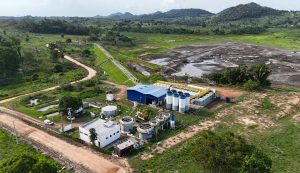 A imagem aérea mostra uma estação de tratamento de água ou esgoto instalada em uma área rural. No primeiro plano, há construções e estruturas industriais, incluindo tanques circulares brancos, reservatórios verticais e um galpão azul. O local é cercado por cercas e cortado por estradas de terra. Atrás da estação, vê-se uma grande área alagada ou de lama escura, possivelmente uma lagoa de decantação, e ao fundo se estende uma paisagem de campos verdes, árvores esparsas e morros sob o céu claro.