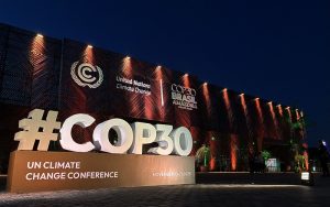 A imagem mostra o local onde será realizada a COP30, a Conferência das Nações Unidas sobre Mudanças Climáticas, marcada para novembro de 2025. É noite, e o céu está escuro, contrastando com a iluminação quente que destaca a fachada do prédio. Em primeiro plano, há uma grande estrutura com a inscrição #COP30 em letras brancas e imponentes, seguida pelas palavras UN Climate Change Conference e as datas November 10–21, 2025. Na parede do edifício, aparecem os logotipos da ONU e do COP30 Brasil Amazônia, indicando que o evento ocorrerá na região amazônica. O ambiente transmite seriedade e importância, sugerindo que se trata de um encontro global sobre o futuro do planeta. A iluminação e os elementos visuais reforçam o caráter oficial e internacional da conferência.