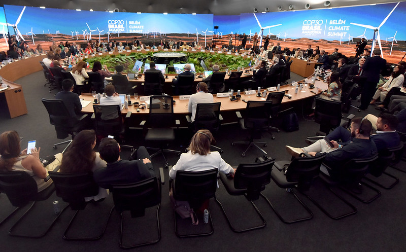 A foto mostra uma grande reunião em formato circular, com dezenas de pessoas sentadas ao redor de uma mesa em forma de anel. No centro há uma área decorada com plantas verdes. Ao fundo, um enorme painel digital exibe a paisagem de um campo com aerogeradores e os dizeres “COP30 Brasil – Belém Climate Summit”. Os participantes, vestidos de forma formal, observam ou conversam entre si, sugerindo uma conferência internacional sobre clima.