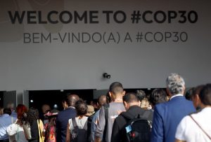 O ChatGPT disse: A foto mostra um grupo de pessoas de costas, caminhando em direção a uma entrada onde há um grande letreiro escrito “WELCOME TO #COP30” e, logo abaixo, “BEM-VINDO(A) A #COP30”. A cena é iluminada pela luz do dia, e o público é diverso, com pessoas vestindo roupas sociais e informais, algumas com crachás de identificação pendurados no pescoço. A imagem transmite o clima de chegada e recepção de participantes ao evento internacional sobre mudanças climáticas.