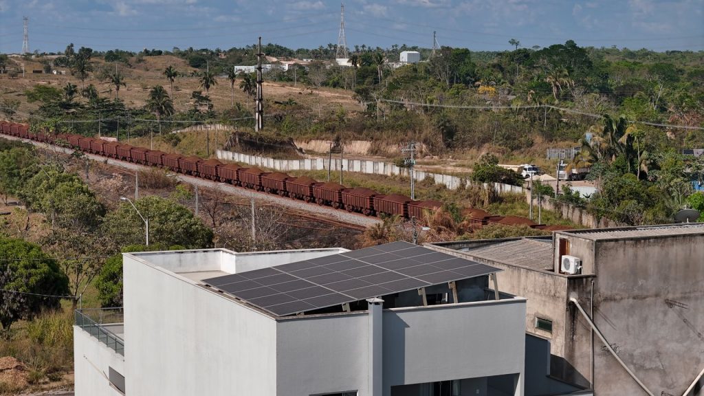 A imagem mostra uma paisagem mista de área urbana e rural. Em primeiro plano, há um prédio de cor clara com painéis solares instalados no telhado. Ao fundo, um trem de carga com vários vagões marrons, cheios de minério, segue por uma ferrovia que atravessa uma região com vegetação esparsa, árvores e morros baixos. Há postes e torres de energia ao longo da linha do trem. O céu está claro e azul, indicando um dia ensolarado.