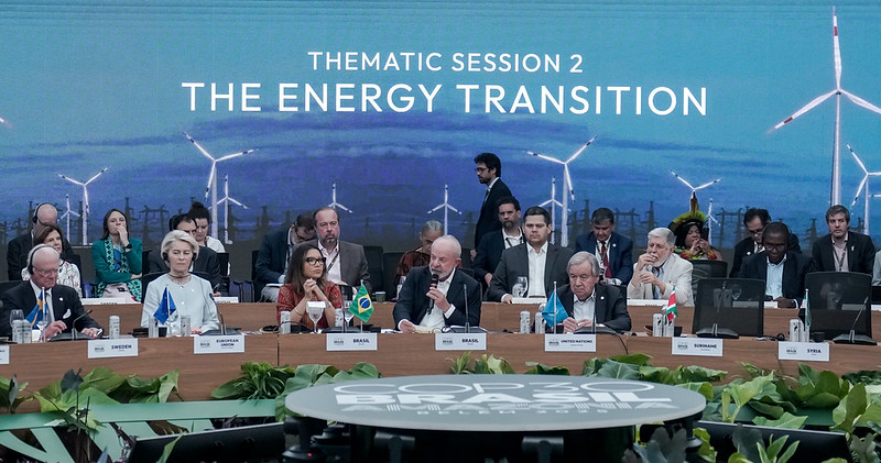 A imagem mostra uma mesa de conferência com autoridades internacionais participando da sessão temática “The Energy Transition” (“A Transição Energética”) durante a COP30. No centro, há o presidente do Brasil falando ao microfone, com a bandeira brasileira à sua frente. Ao redor dele, estão representantes de diversos países, cada um identificado por uma pequena placa e bandeira nacional. O fundo do palco exibe uma imagem ampliada de turbinas eólicas, simbolizando energia limpa. O ambiente é formal, com iluminação clara e decoração com plantas sobre a mesa principal.